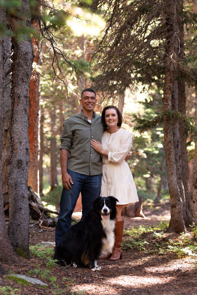 A couple smile for their engagement photo with their dog in the woods at Brainard Lake