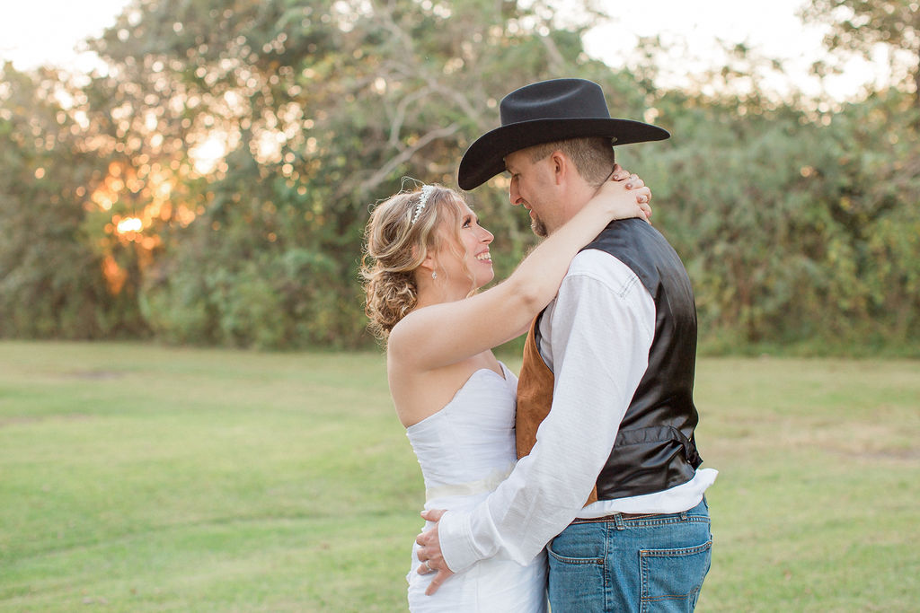 A groom in a cowboy hat embraces his bride in a lawn at sunset