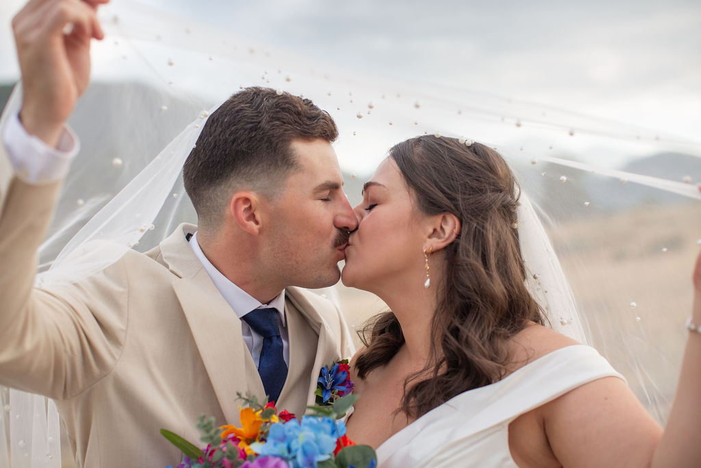 Newlyweds kiss while hiding under and holding up the veil at sunset with pearls