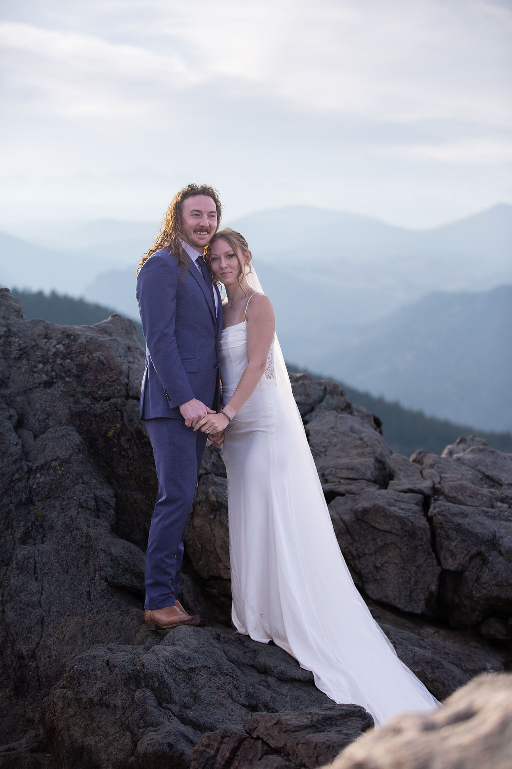 A bride leans into the chest of her groom as they hold hands in the mountains on boulders