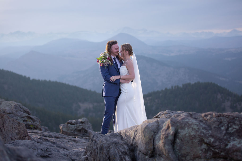 A bride and groom embrace on a mountaintop at sunset and lean in for a kiss while eloping in boulder