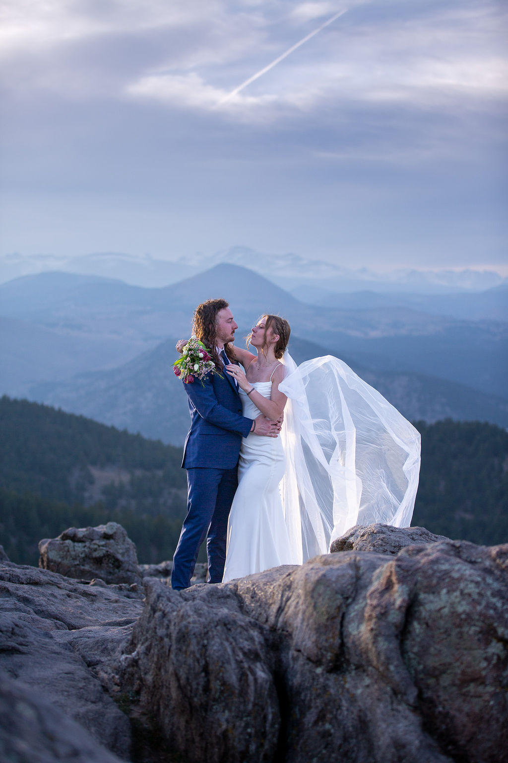 Newlyweds snuggle while standing on a mountain overlook at sunset as the veil flies in the wind while eloping in boulder