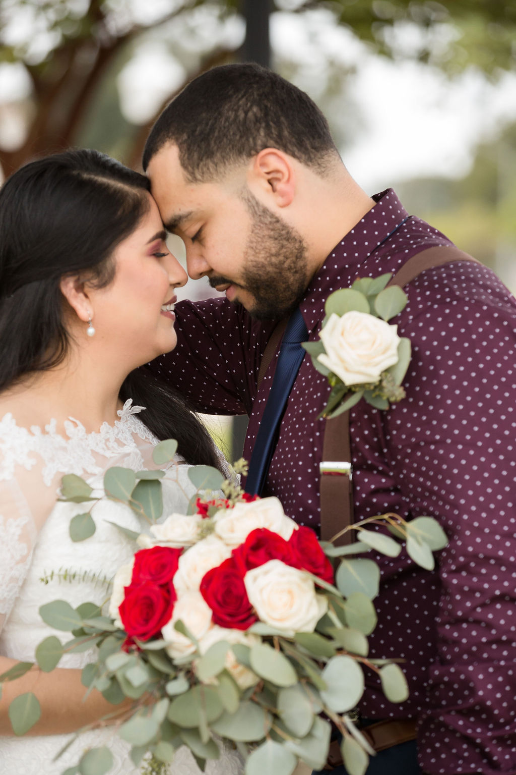 A bride smiles while leaning in for a kiss with her groom in a purple shirt and suspenders