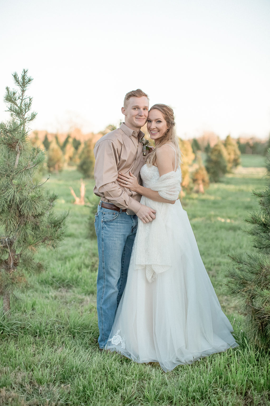 A bride and groom snuggle in a christmas tree farm at sunset