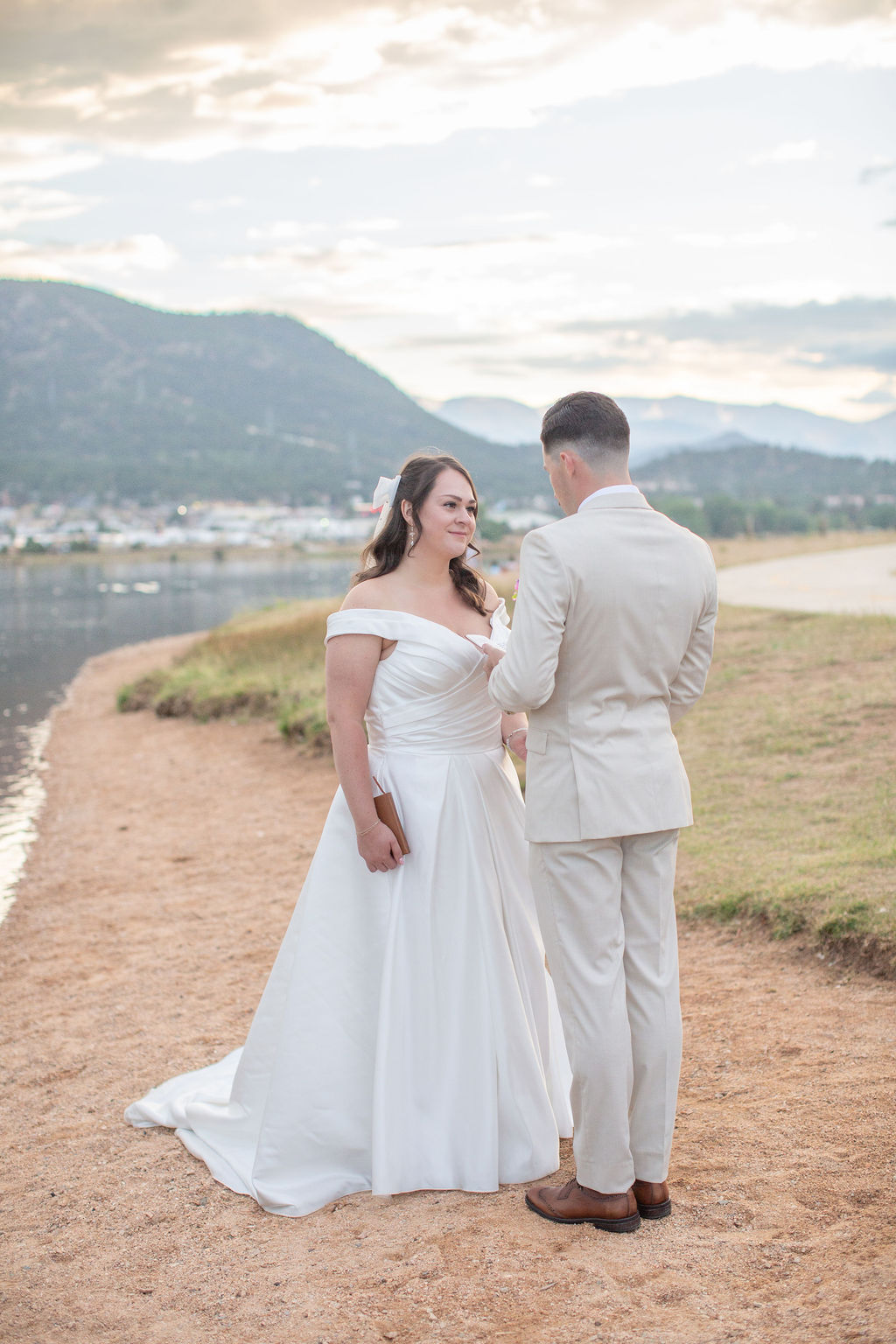 A groom in a tan suit reads his vows to his bride on the lake's edge at sunset