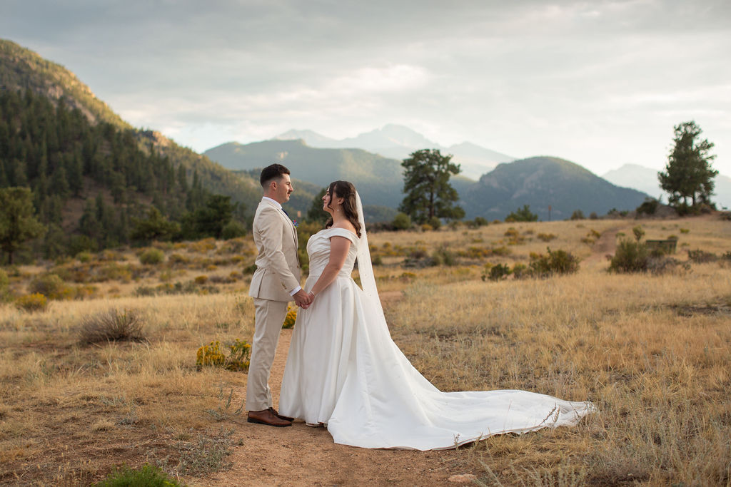A groom in a tan suit stands holding hands and facing his bride in an alpine trail at sunset with smiles during their rocky mountain national park elopement