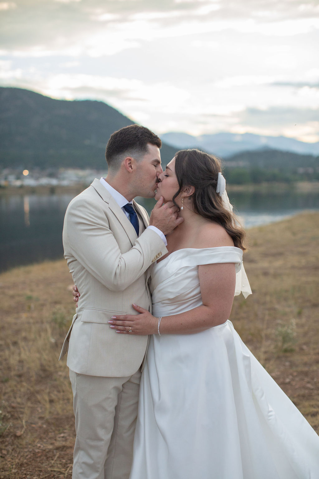 Newlyweds kiss by the water at sunset in a tan suit and silk dress during their rocky mountain national park elopement