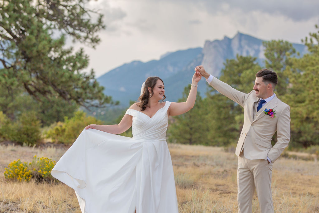 A groom in a tan suit holds the hand of his bride as she twirls in a meadow with wildflowers near the mountains at their taharaa mountain lodge wedding