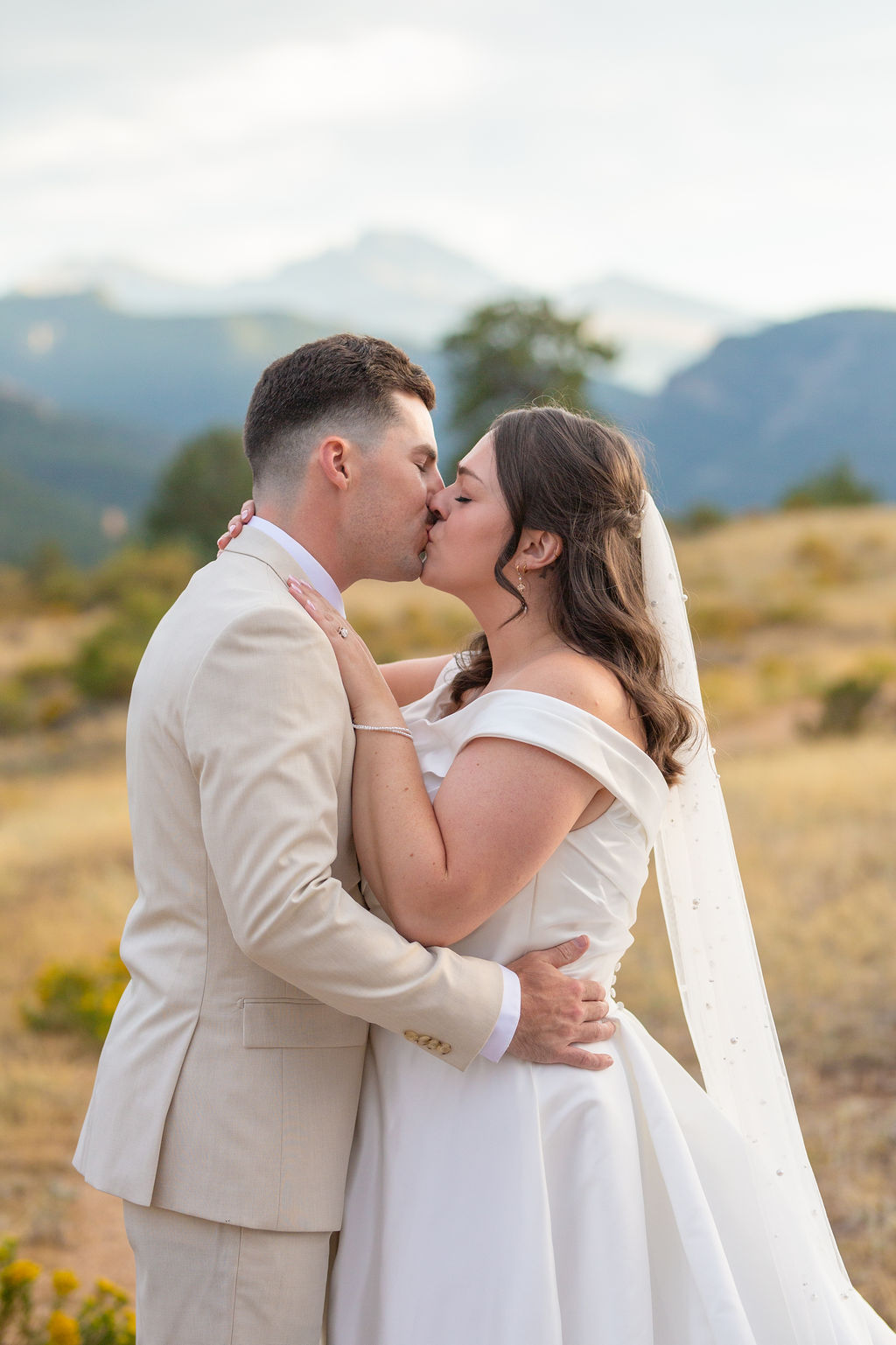 Newlyweds kiss on a hillside during their taharaa mountain lodge wedding in a tan suit and silk gown