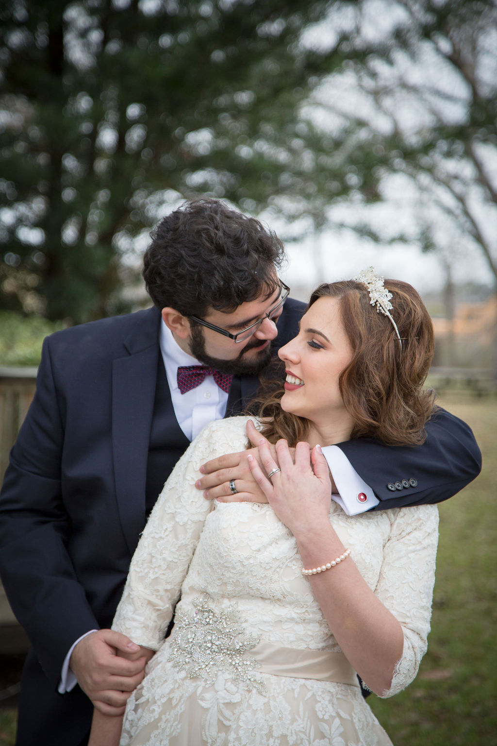 A bride and groom snuggle and lean in for a kiss in a lawn under trees at their yellow barn farm wedding