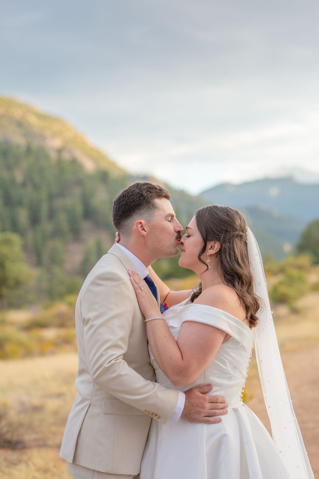 A groom in a tan suit kisses the nose of hims bride at sunset in the mountains during an estes park elopement
