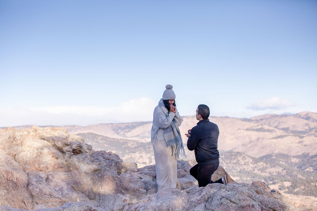 A man in black kneels during a mountain hike to propose to his ecstatic girlfriend in a scarf and beanie at one of the epic estes park elopement locations