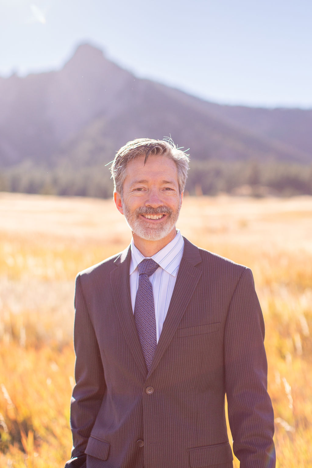 A groom in a stripe suit stands in a meadow of tall golden grass at sunset during his intimate wedding in colorado