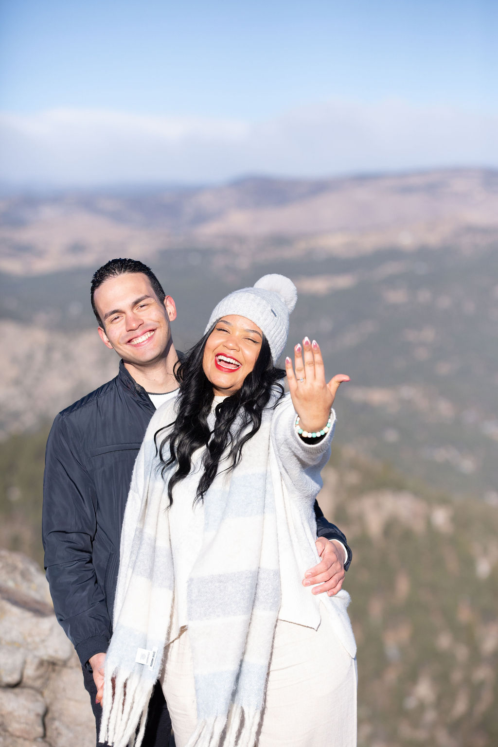 Happy engaged woman shows off her ring on a mountain overlook as her fiancee smiles and hugs her before finding a micro-wedding planner colorado