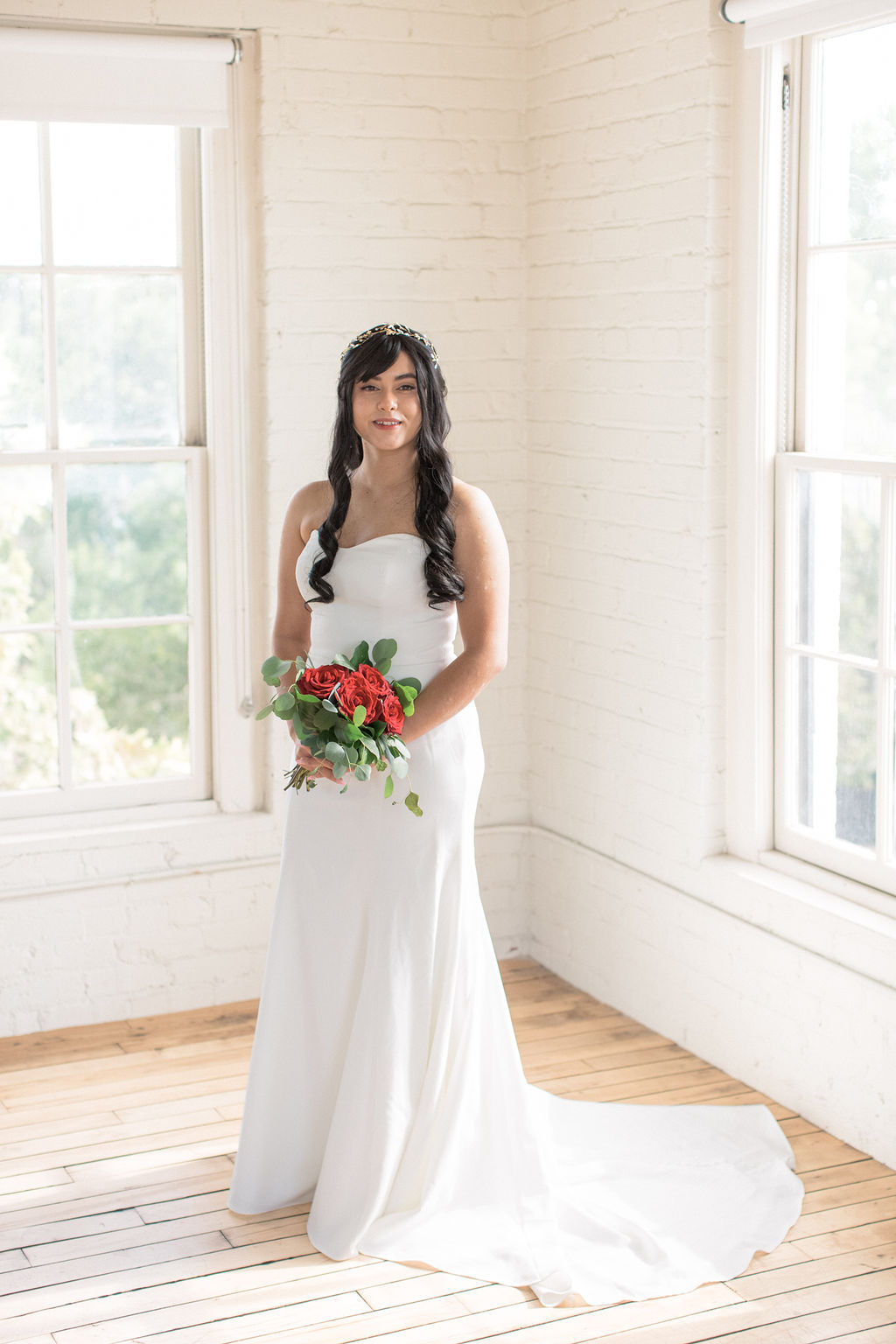 A bride stands in a corner of a white brick room full of windows holding her red rose bouquet