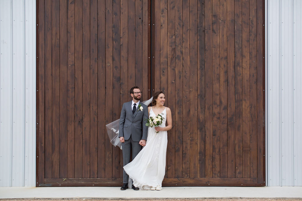 A bride and groom stand against large wooden doors holding hands and looking to the side