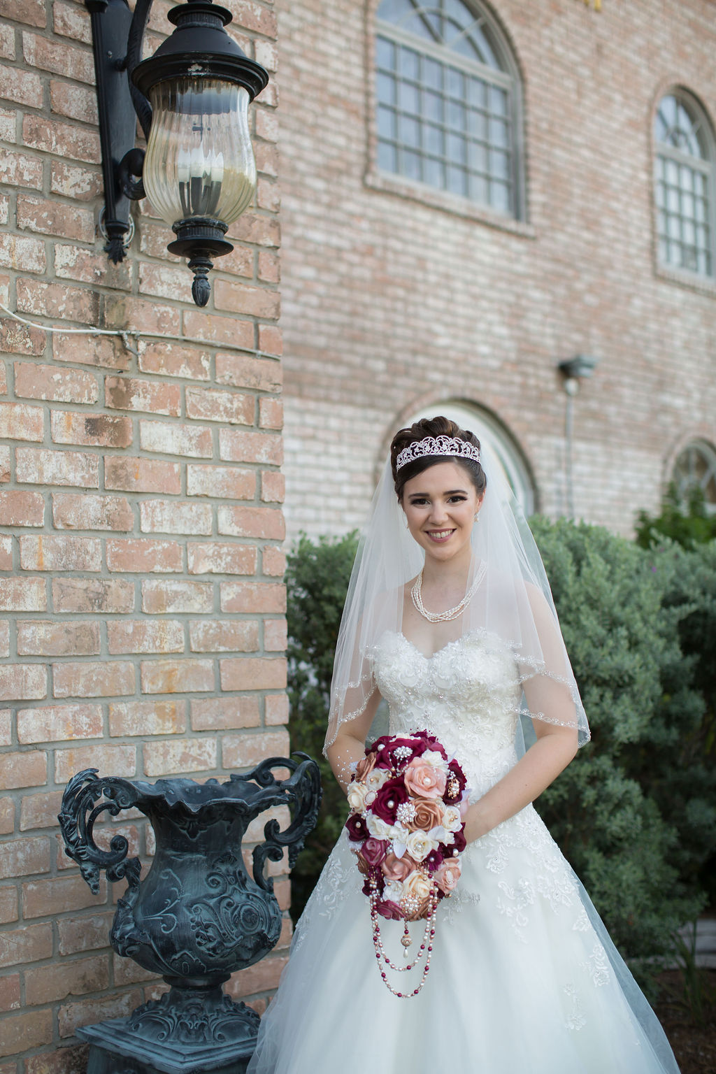 A happy bride stands by an exterior light in a garden by a brick building smiling and holding her maroon bouquet