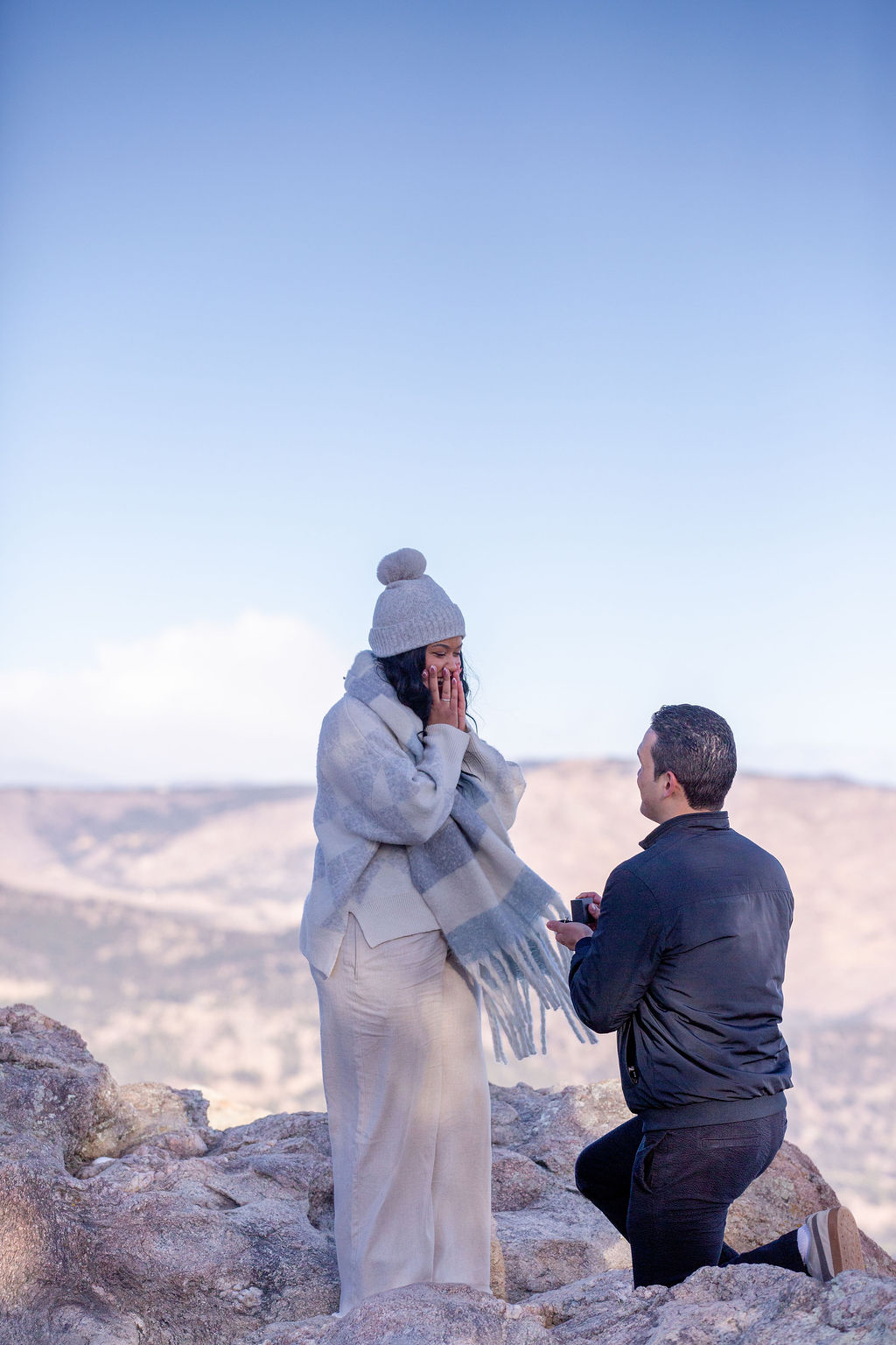 A man on one knee proposes to his now fiancee in the montains during a hike in the cold