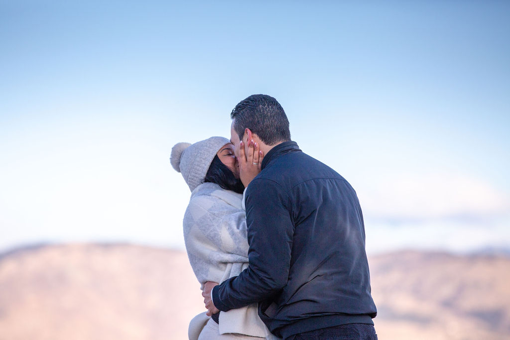Newly engaged couple kisses on a mountaintop in grey and white jackets before exploring wedding planners in boulder