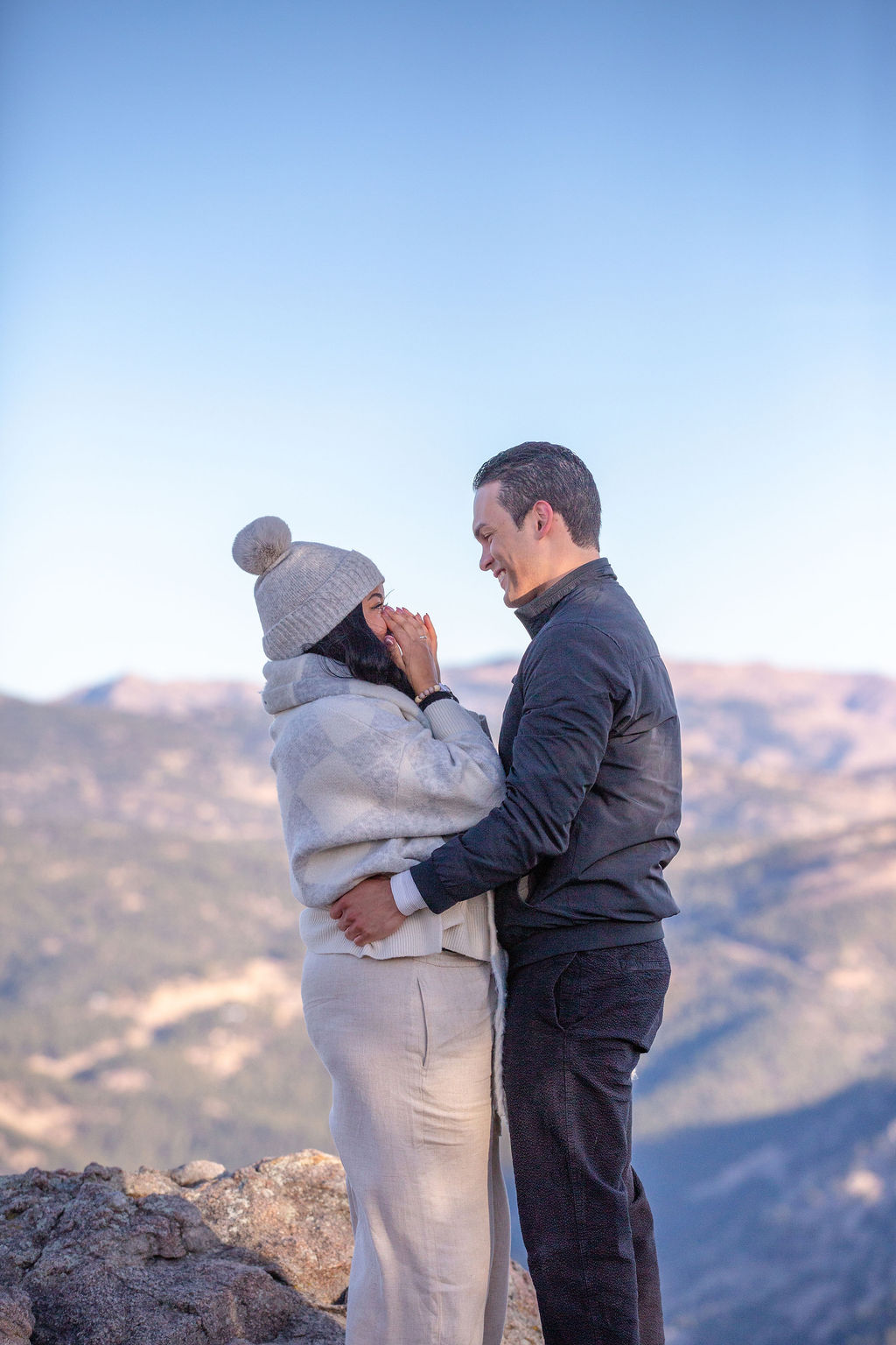 A woman covers her face in joy after her proposal on a mountain overlook while her fiancee hugs her before finding wedding planners in boulder