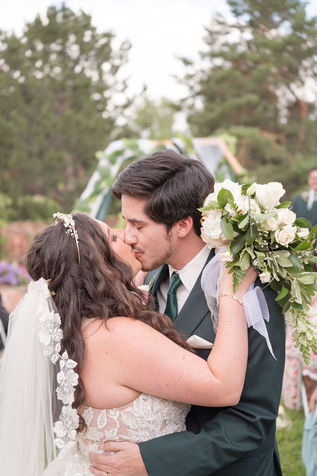 Newlyweds kiss in the aisle in a green suit and lace floral gown as guests watch