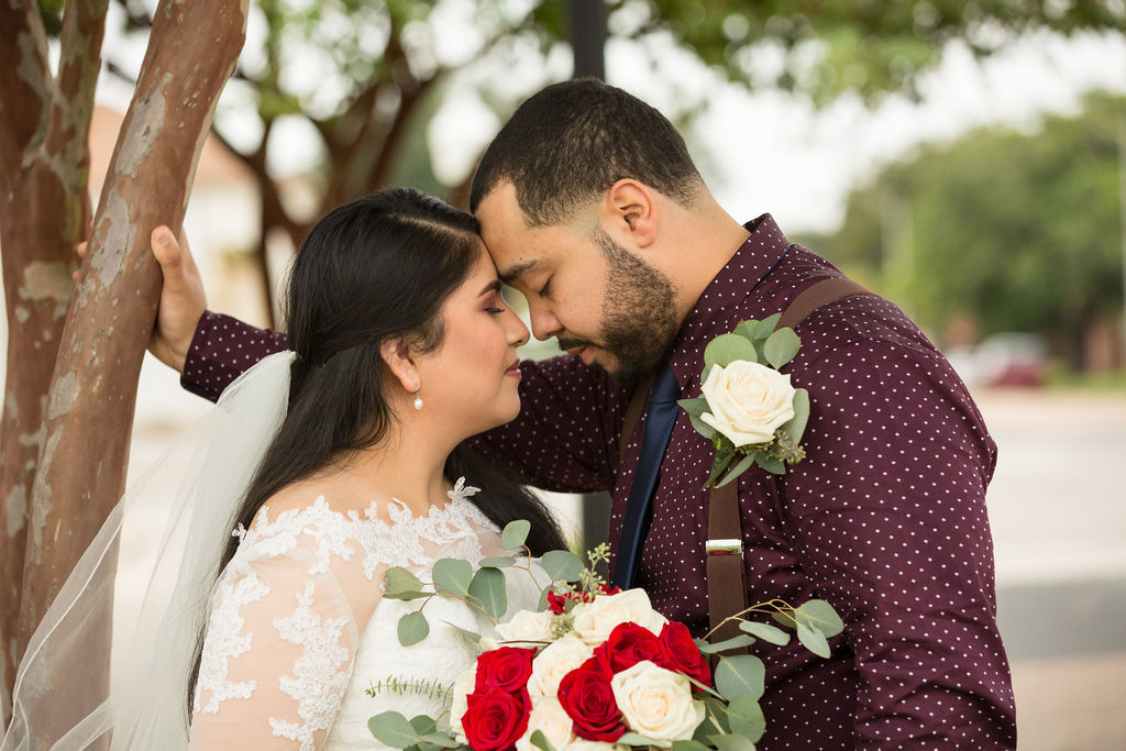 A groom in maroon shirt and suspenders leans over his bride as they touch foreheads against a tree after using wedding planners in estes park, CO