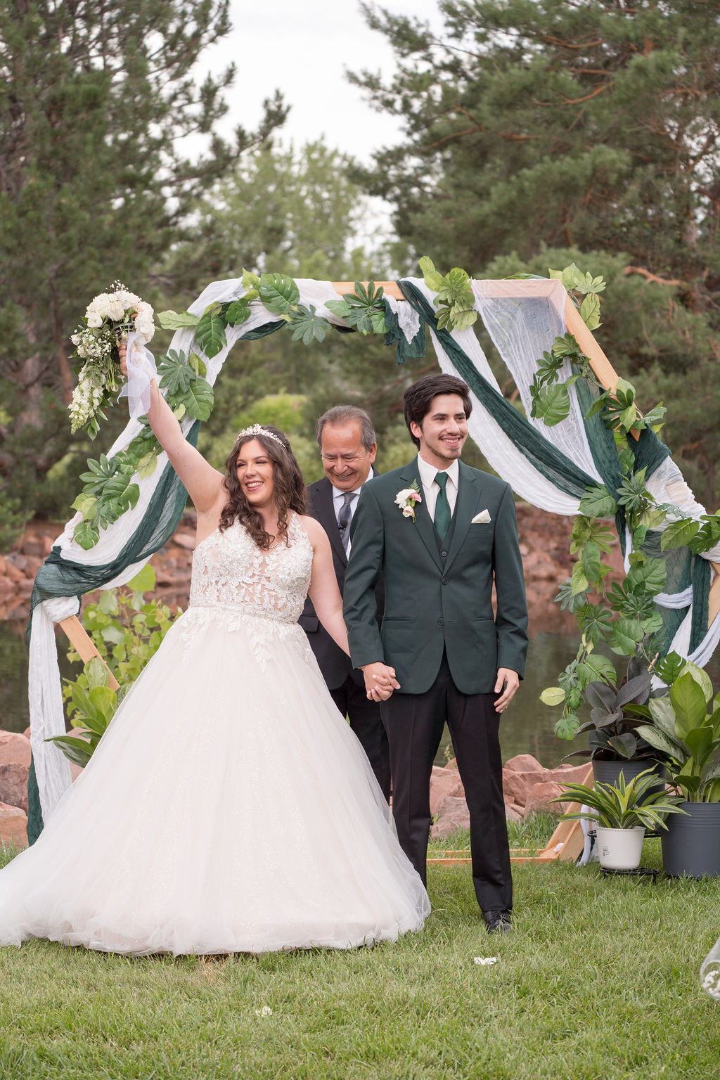 A bride lifts her bouquet at the outdoor altar holding her groom's hand in a green suit at one of the wedding venues in lyons co
