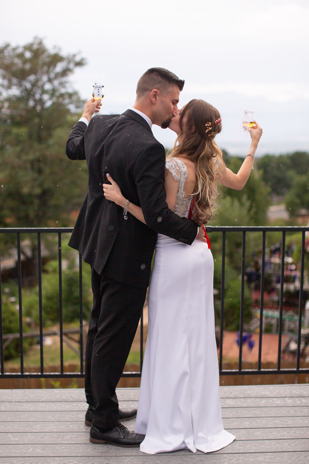 Newlyweds lift champagne glasses and kiss on a balcony with arms around each other