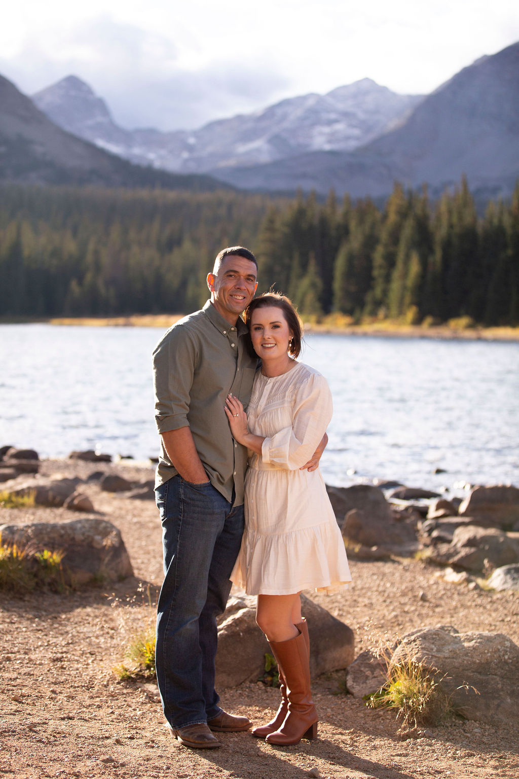 Happy couple snuggle in front of an alpine lake at sunset in white and green after exploring wedding venues in northern colorado